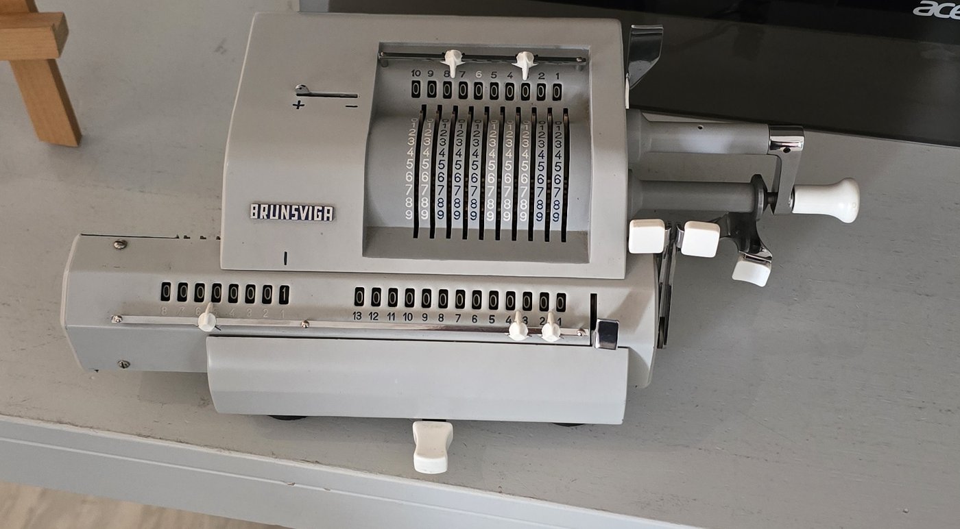A grey Brunsviga mechanical calculator with number wheels, setting levers and a hand crank, on a museum table