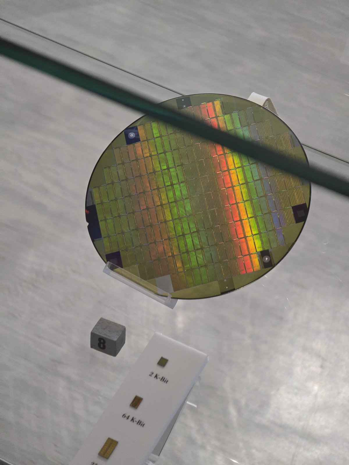 A silicon wafer of memory chips in a display case, with rainbow colours across it, above labelled single chips of 2 K-bit and 64 K-bit